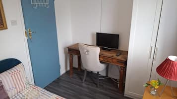 Una habitación con una puerta azul, un escritorio de madera con una computadora, una silla blanca, una cama con ropa de cama estampada y una lámpara roja sobre una mesita de noche.
