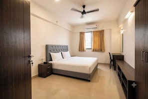 A bedroom with a bed, bedside table, wardrobe, ceiling fan, and window with curtains. - Townhouse Nanded City (Pune)
