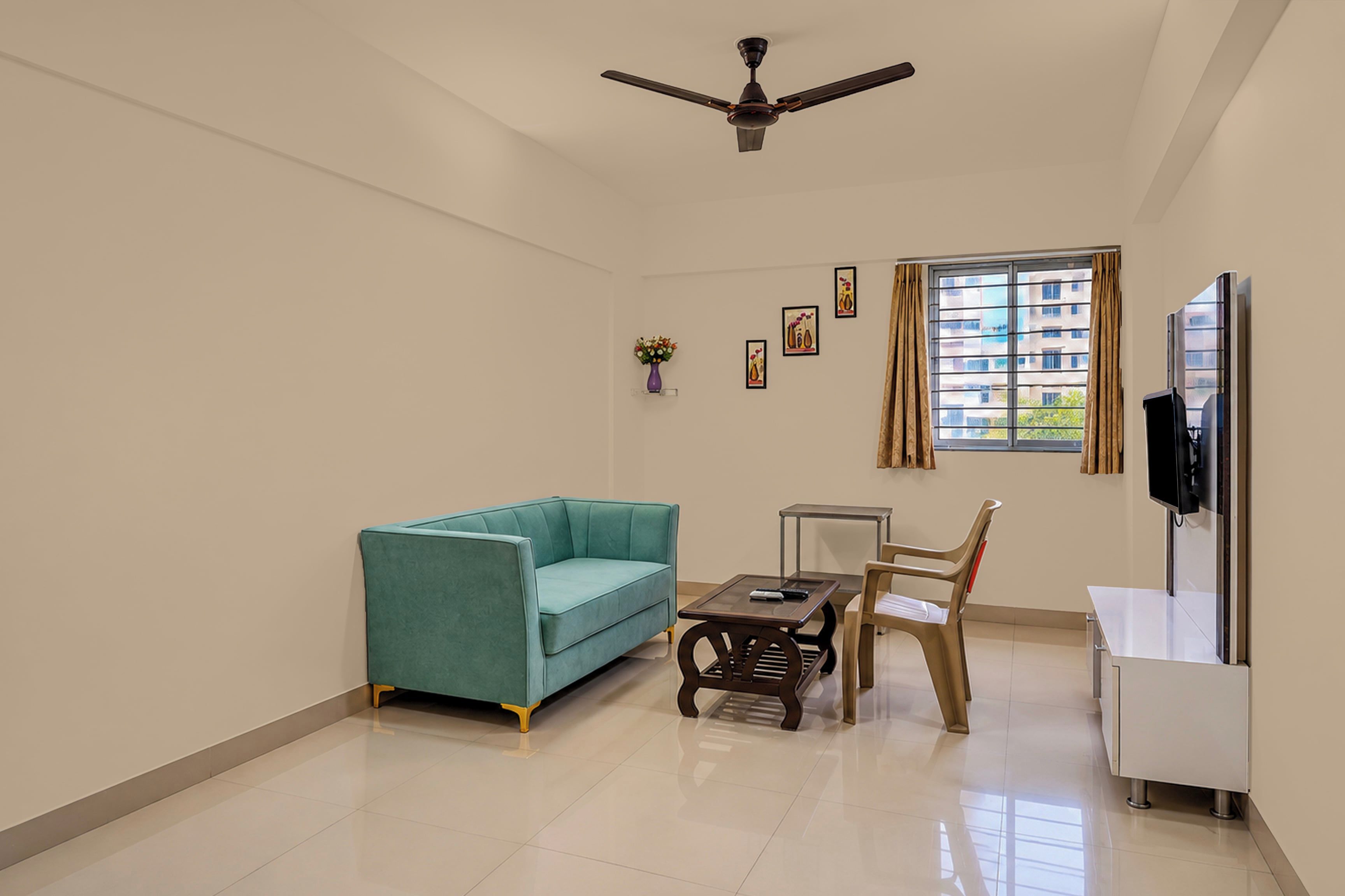 Una habitación con un sofá color turquesa, una mesita, una silla, un ventilador de techo, una ventana con cortinas y un televisor montado en la pared.
