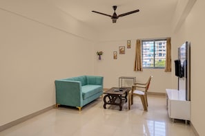 A room with a teal sofa, a small table, a chair, a ceiling fan, a window with curtains, and a television mounted on the wall.