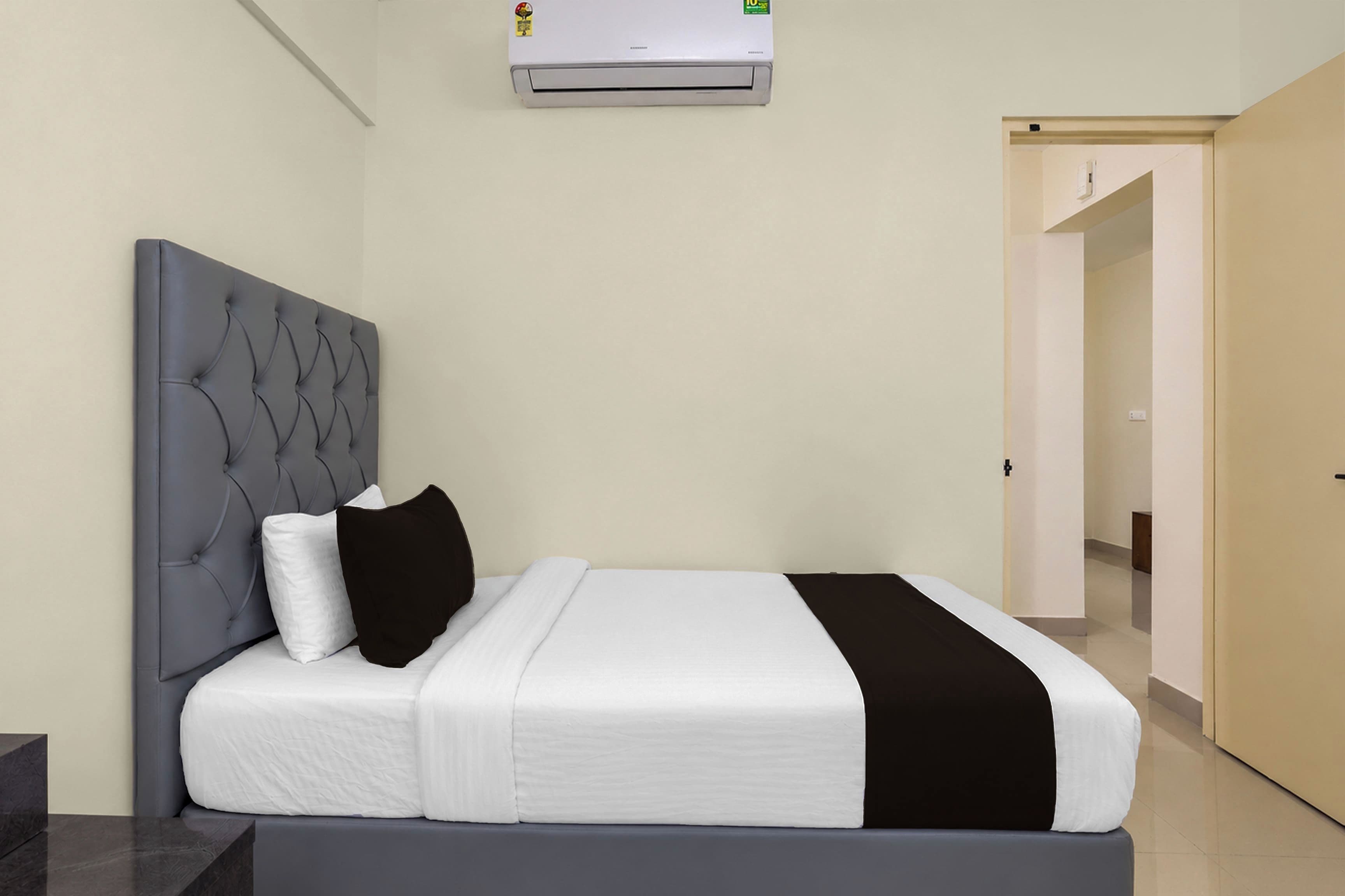 Een hotelkamer met een bed, een grijs bekleden hoofdeinde, wit beddengoed, een zwarte plaid en een airconditioner boven het bed.
