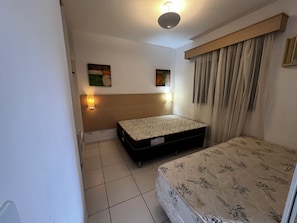A small, clean room with a bed, a nightstand, a wall-mounted lamp, and a window with curtains. - CRCN - Pontal do Lago (Caldas Novas)