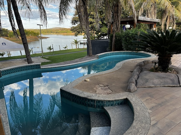 A swimming pool with a curved design, surrounded by palm trees and a view of a lake. - CRCN - Pontal do Lago (Caldas Novas)