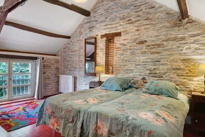 Une chambre avec un mur en pierre, un lit avec une literie à motifs floraux, une fenêtre avec des rideaux et un radiateur.