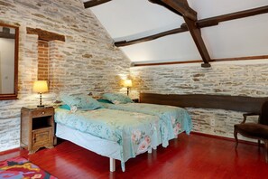 A bedroom with a stone wall, wooden beams, a bed with floral bedding, a nightstand, a chair, and a rug. - Apartment 'Gîte La Rosa - Domaine Les Bois De' with Shared Pool, Shared Terrace and Wi-Fi (Sixt-sur-Aff)