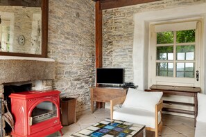 Una habitación rústica con pared de piedra, un horno de leña rojo, un televisor sobre una mesa de madera y una silla blanca.