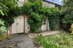 A rustic stone building with a wooden door and a small window, surrounded by lush greenery and a wooden pathway. - Apartment 'Gîte La Rosa - Domaine Les Bois De' with Shared Pool, Shared Terrace and Wi-Fi (Sixt-sur-Aff)