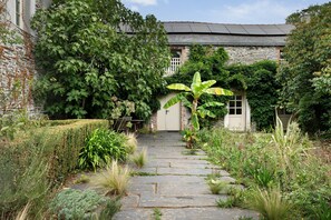 A stone pathway leading to a building with a white door, surrounded by lush greenery and a brick wall. - Apartment 'Gîte La Rosa - Domaine Les Bois De' with Shared Pool, Shared Terrace and Wi-Fi (Sixt-sur-Aff)