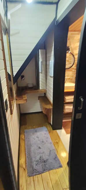 Um corredor estreito com piso de madeira, um pequeno degrau dando acesso a um nível mais alto, e uma porta à direita.