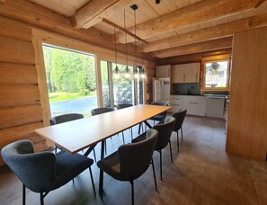 Una cocina moderna con vigas de madera, un comedor con una mesa larga y sillas, y vista a un paisaje verde a través de grandes ventanales.