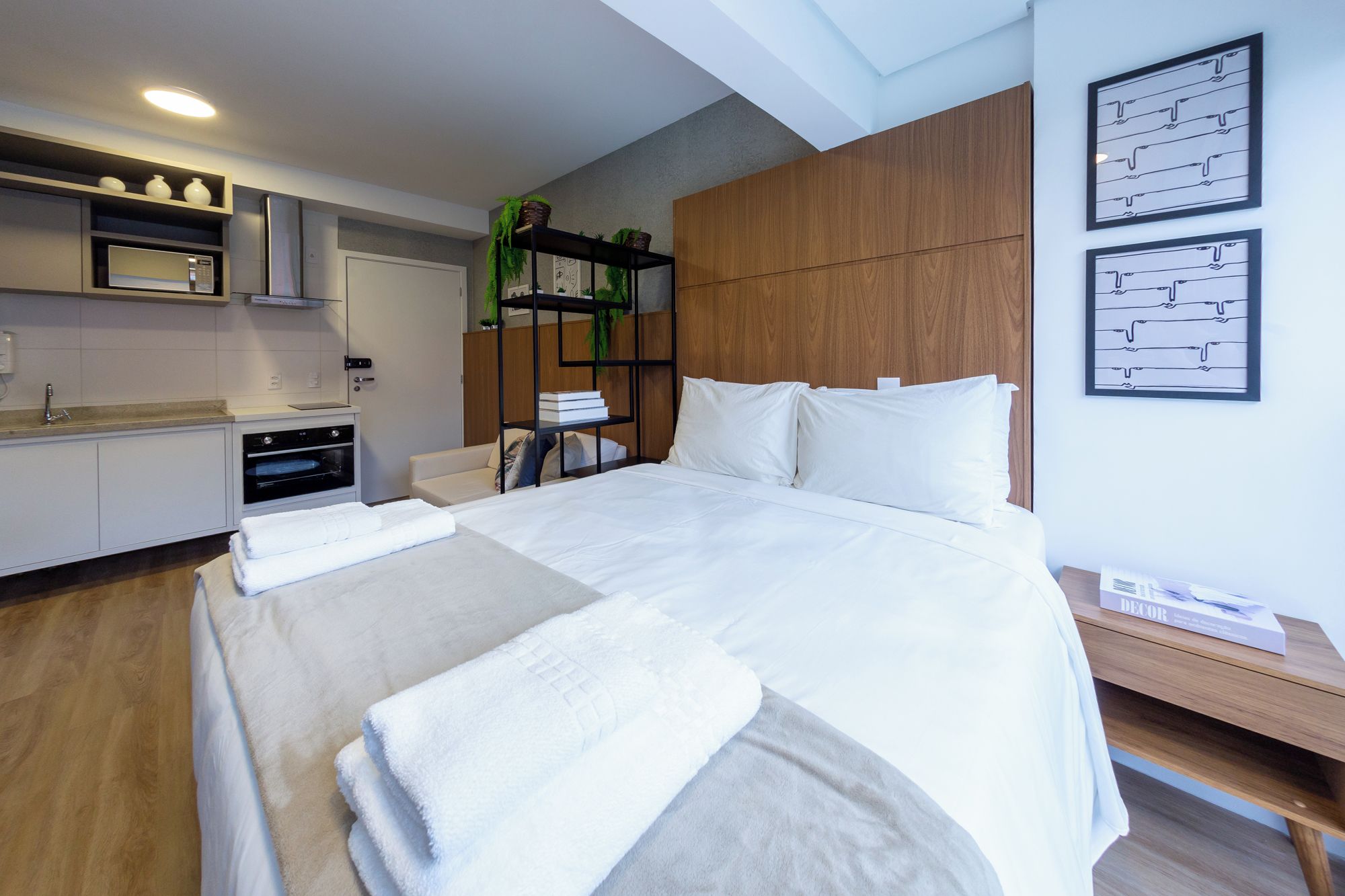 Ein modernes Hotelzimmer mit einem Bett, einem hölzernen Kopfteil, einer eingebauten Ablage mit Pflanzen, einer kleinen Küchenzeile und einem Nachttisch mit einem Buch.