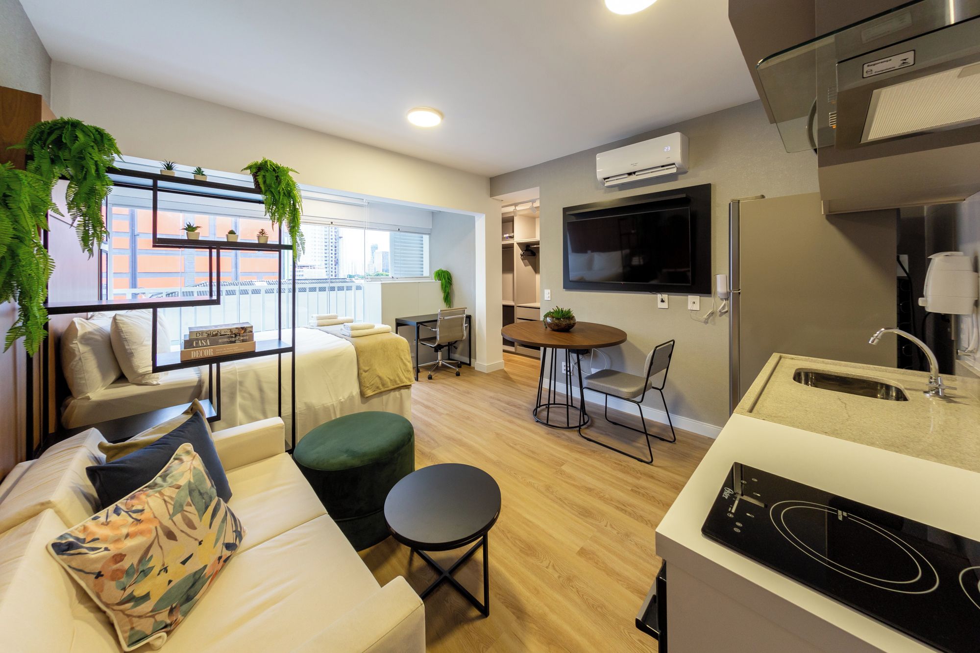 Een modern appartement met kitchenette, een eethoek, een zithoek met een bank en voetenbankje, en een groot raam met uitzicht op de stad.