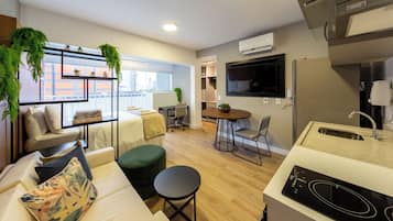 Een modern appartement met kitchenette, een eethoek, een zithoek met een bank en voetenbankje, en een groot raam met uitzicht op de stad.