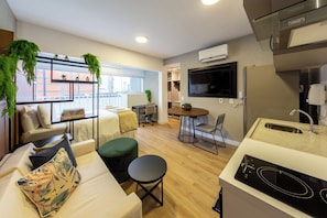 Een modern appartement met kitchenette, een eethoek, een zithoek met een bank en voetenbankje, en een groot raam met uitzicht op de stad.