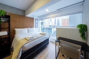 A modern hotel room with a bed, a desk, a chair, a bookshelf, and a large window with a city view. - Just by Bhomy (São Paulo)