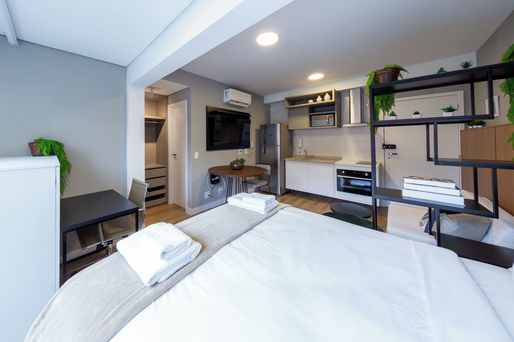 A compact hotel room with a bed, a desk, a kitchenette, and a TV.