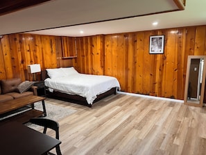 A bedroom with a wooden paneled wall, a bed, a nightstand, a lamp, a wooden bench, and a door.