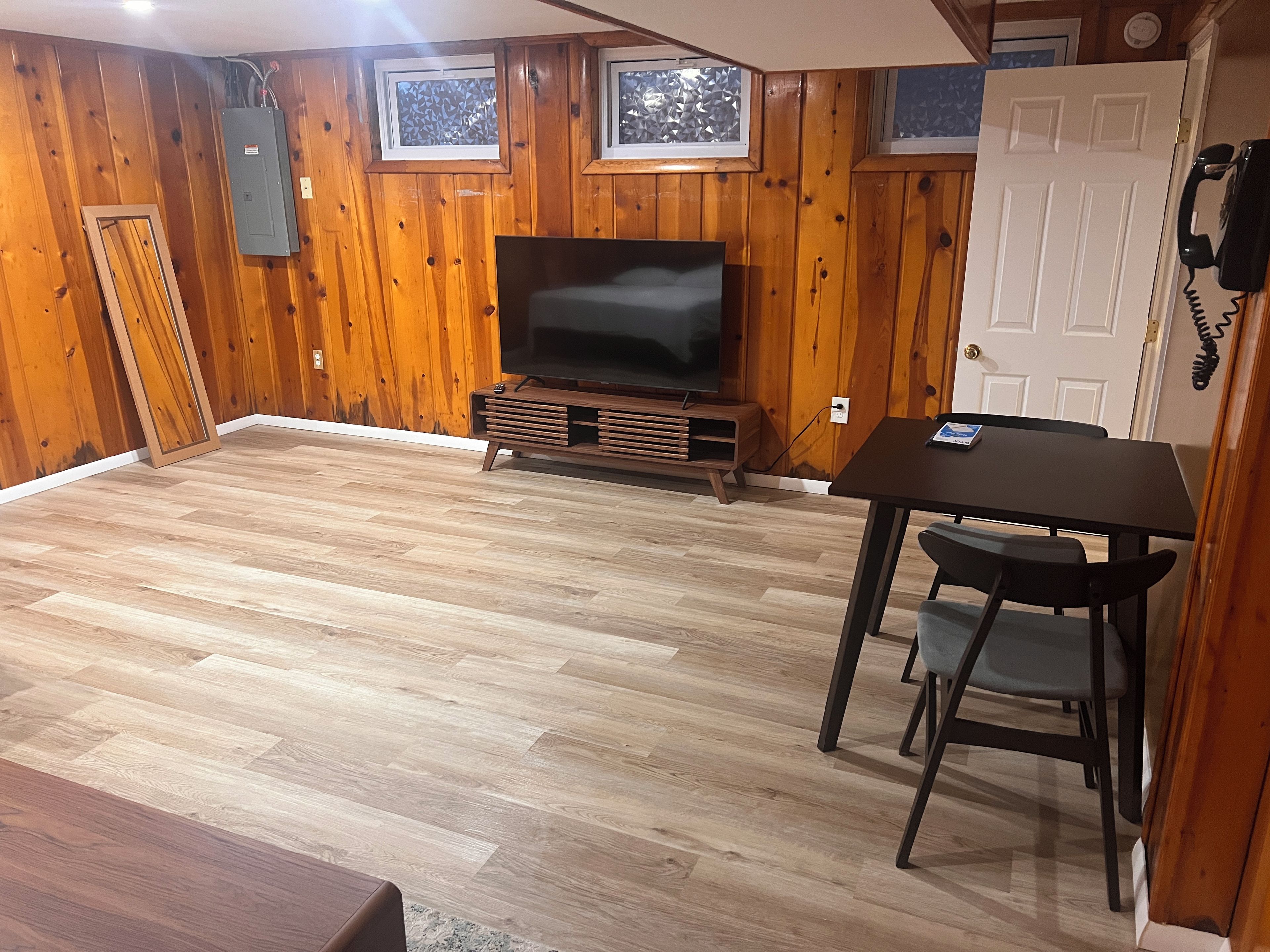 Quarto com paredes de madeira, uma televisão de tela plana sobre um suporte de madeira, uma mesa de jantar com cadeiras e uma porta com telefone.