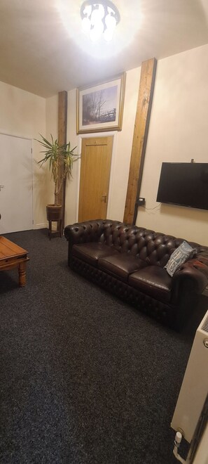 A living room with a dark brown leather sofa, a wooden coffee table, a potted plant, a wooden door, and a flat-screen TV mounted on the wall. - Greystones nawab sleep 25 peak park ..Derbyshire dales..buxton (Buxton)
