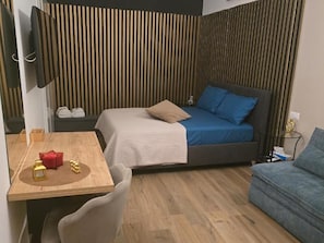Un dormitorio con cama, mesita de noche, una silla, televisor y piso de madera.