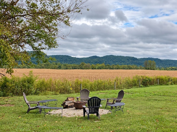 An outdoor seating area with chairs and a fire pit, surrounded by a grassy field and a view of a forested hill.