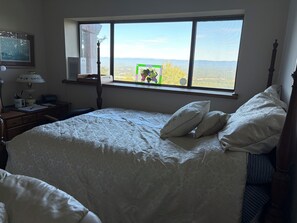 Ein Schlafzimmer mit einem großen Bett, einem Fenster mit Blick auf die Landschaft, einem Schreibtisch mit Lampe und einem gerahmten Bild an der Wand.