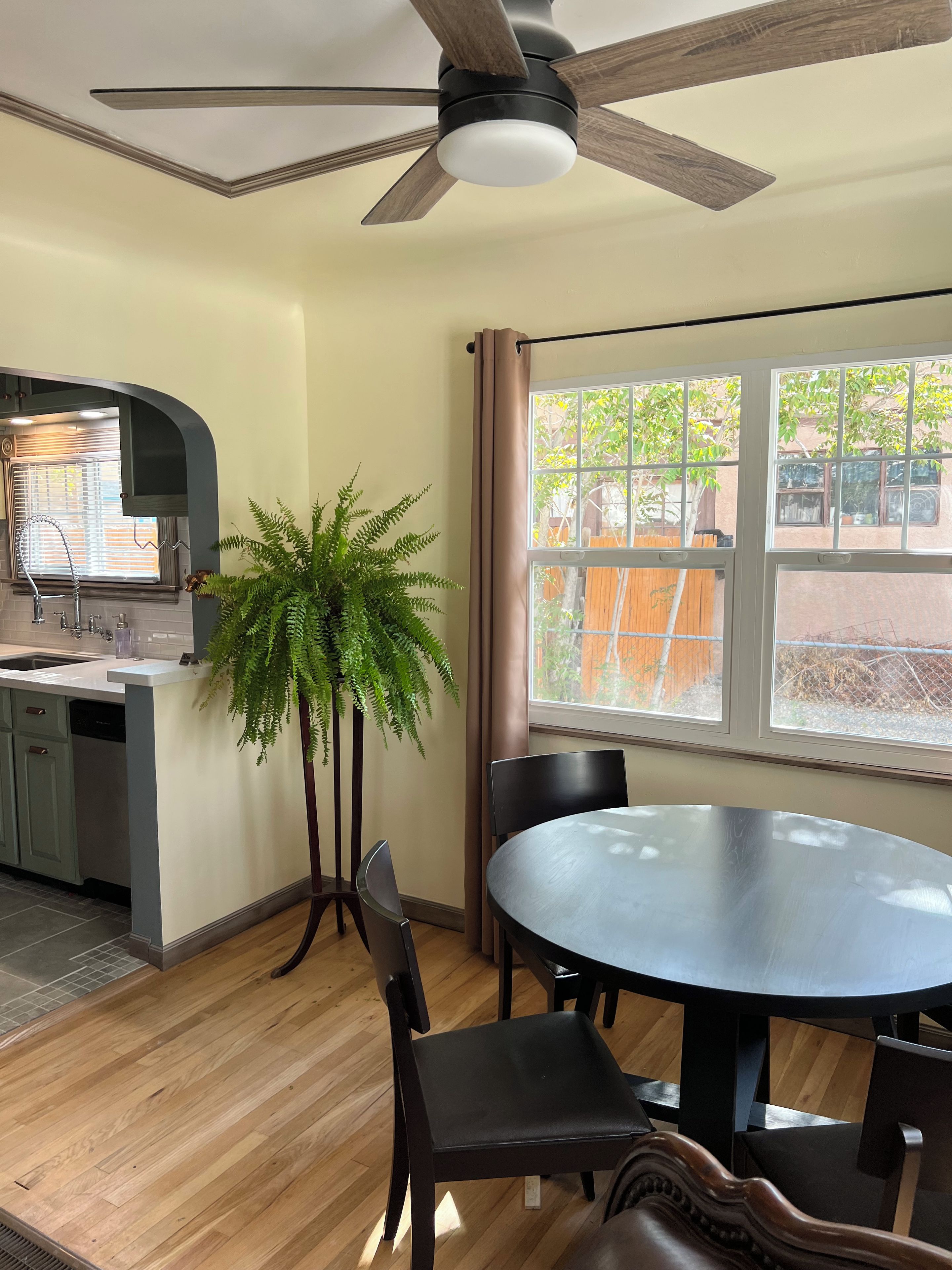 A dining area with a round table, chairs, a ceiling fan, a window with curtains, and a kitchen area with a sink and faucet.