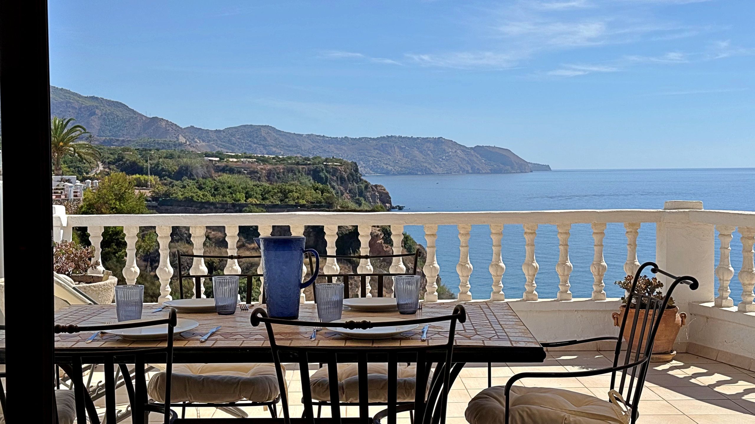 An outdoor dining area with a table set for two, overlooking a coastal landscape with mountains and the sea.