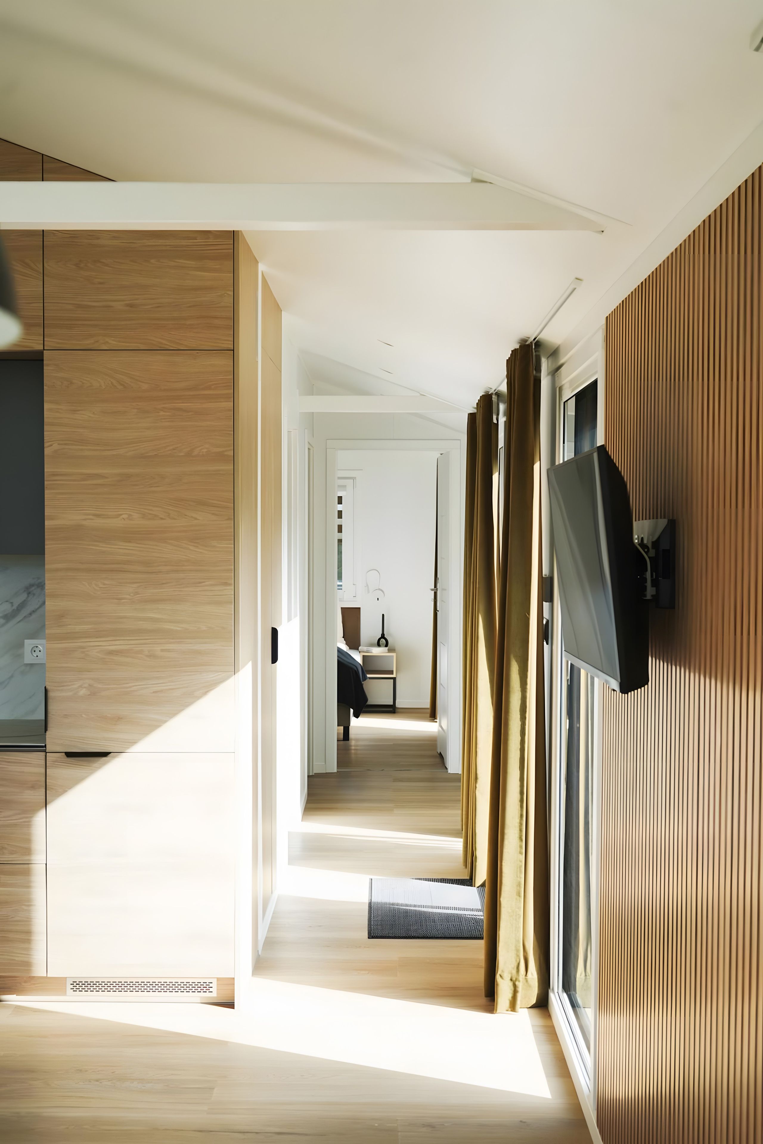 A modern interior with a wooden floor, a flat-screen TV mounted on the wall, and a view into a bedroom with a bed and a desk.