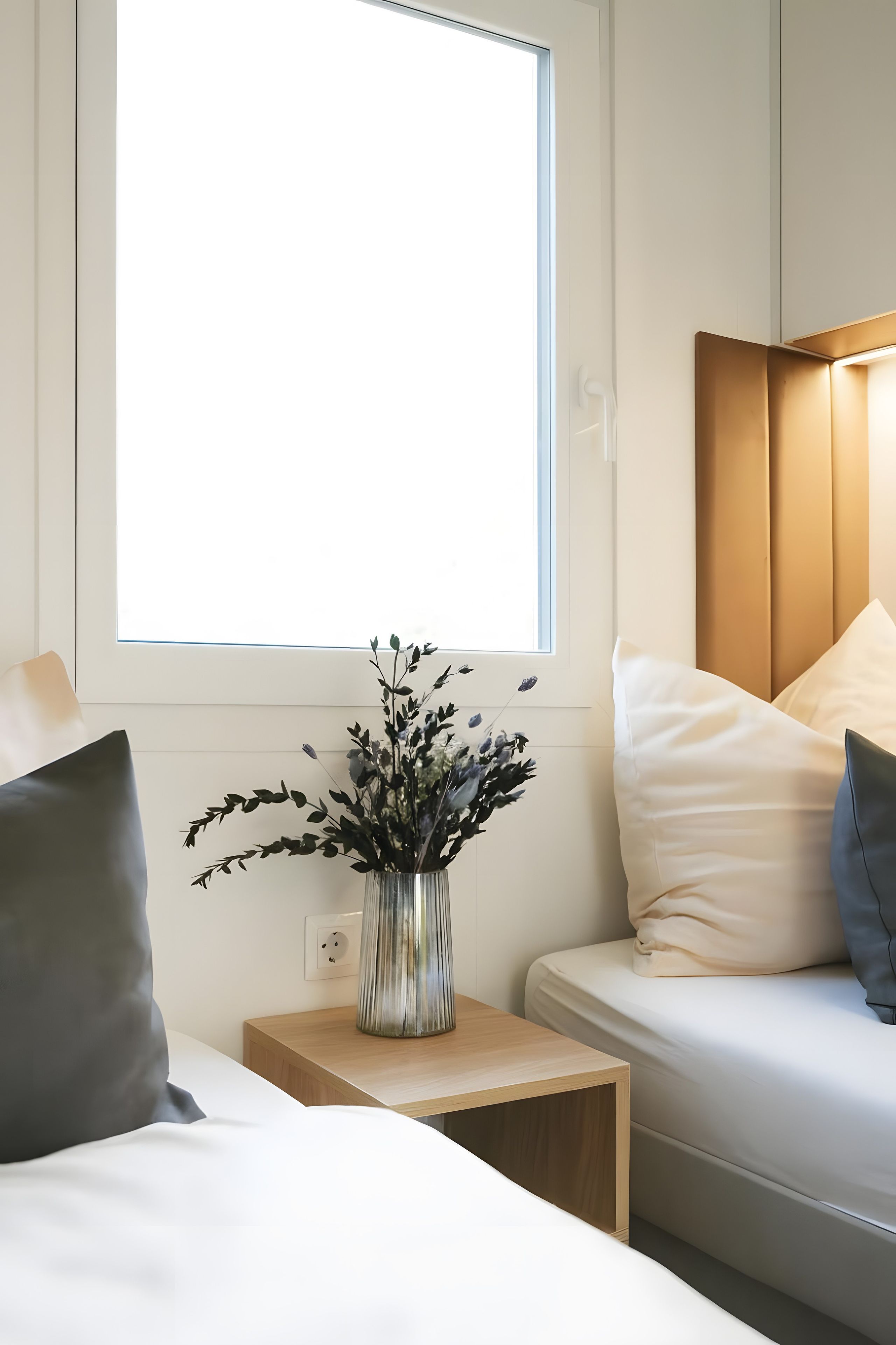 Una camera d'albergo con un letto, un comodino con un vaso di fiori, una finestra con persiane e una testiera con illuminazione integrata.
