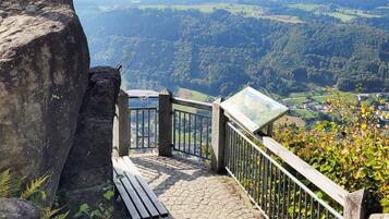 Un sentier de pierres mène à une plateforme d’observation dotée d’une rambarde en métal, d’où l’on peut admirer une vallée verdoyante et des bâtiments situés au loin.