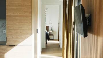 A modern interior with a wooden floor, a flat-screen TV mounted on a wall, and a view into a bedroom through a doorway.
