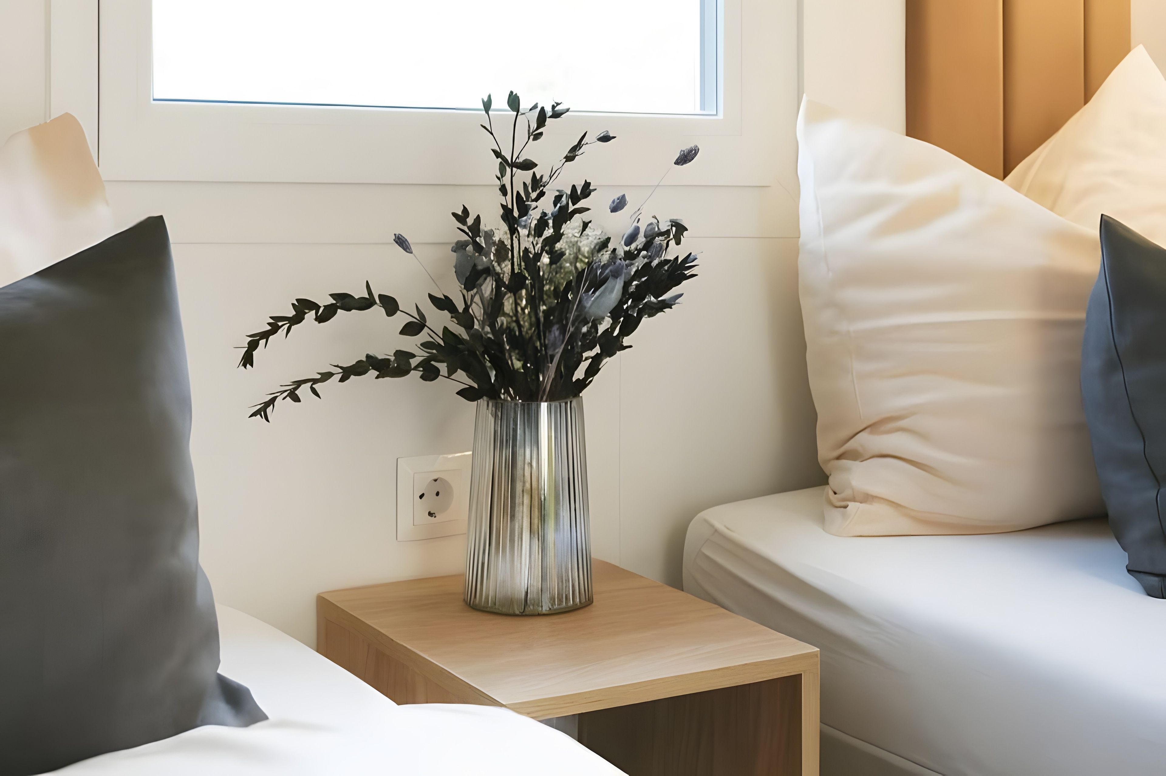 Een slaapkamer met nachtkastje, bloemenvaas, bed met kussens en raam.