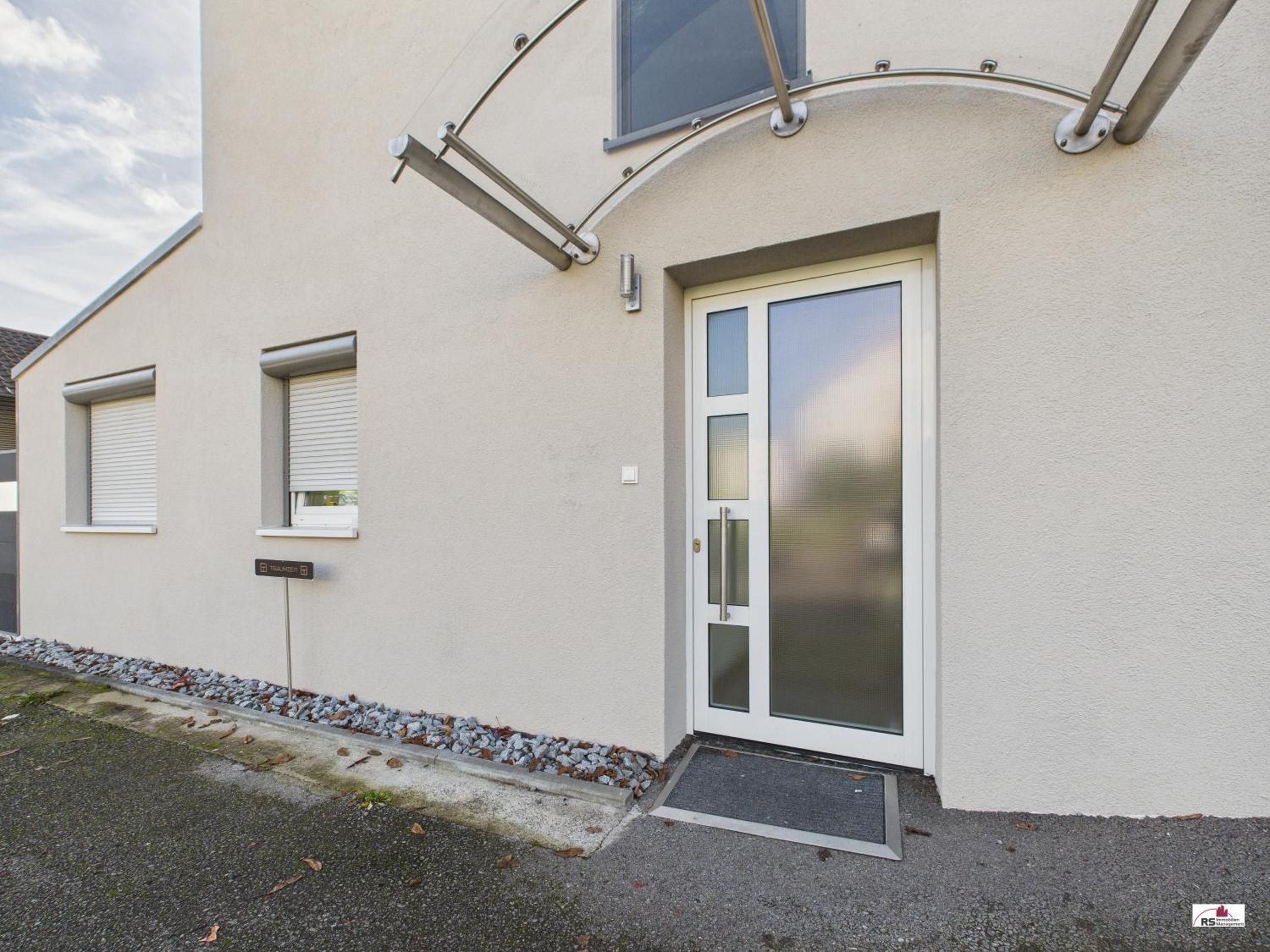 Ein modernes Gebäude mit weißer Fassade, einer Glastür mit Metallgriff und einem kleinen Eingangsbereich mit Fußmatte.