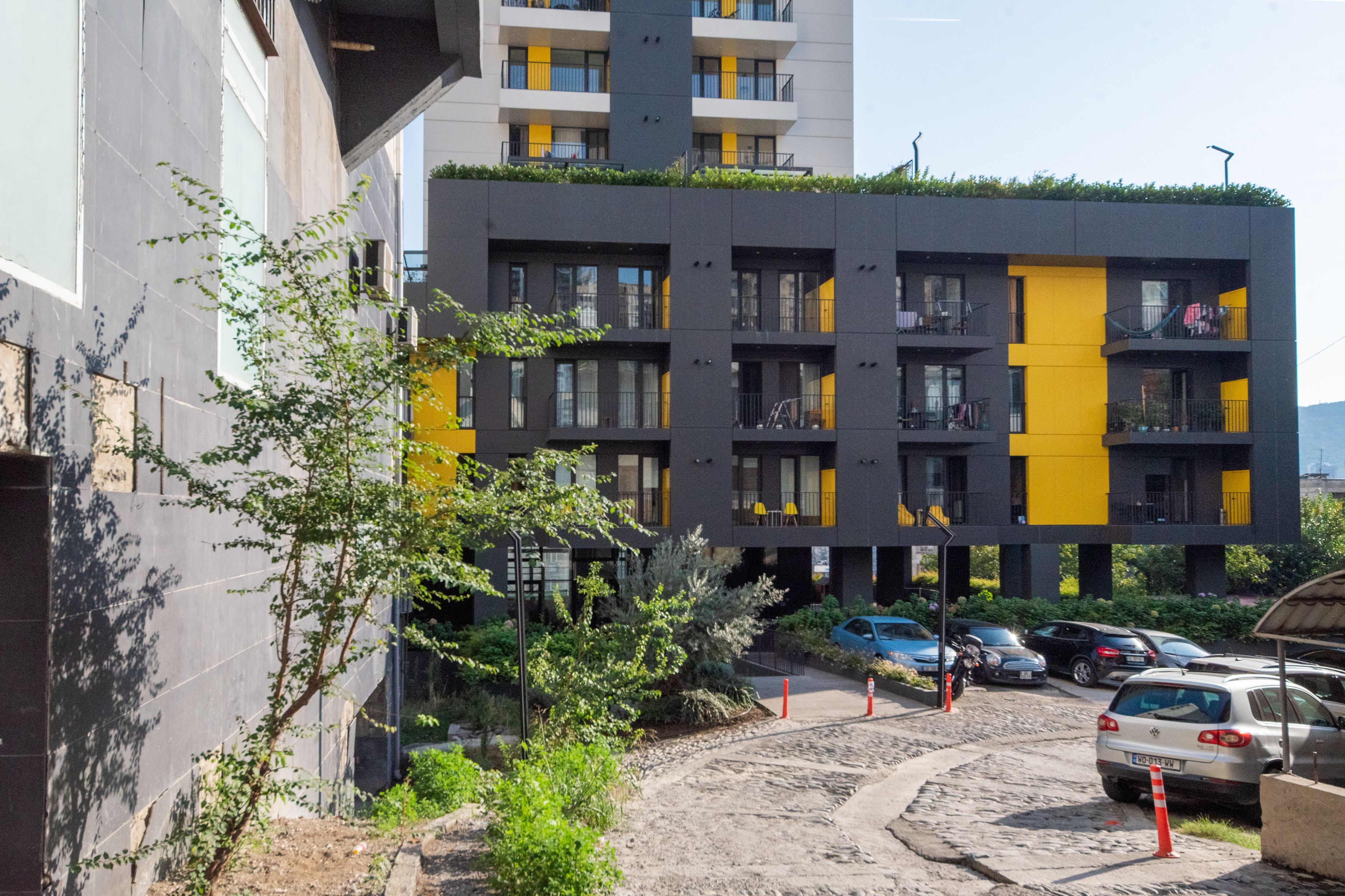 A modern multi-story building with balconies, a parking area, and a dirt path.