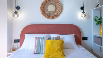 A bed with white bedding, a red headboard, and multiple pillows including a yellow one. There is a wall-mounted mirror above the bed and two wall-mounted lights on either side.