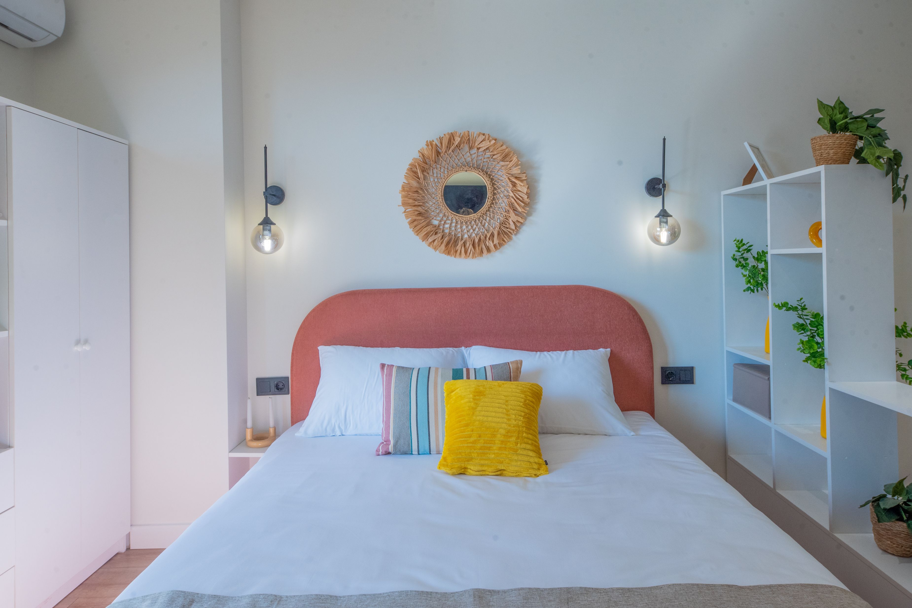 A bedroom with a bed, a headboard, a wall-mounted mirror, two wall sconces, a bedside table, and a white wardrobe.