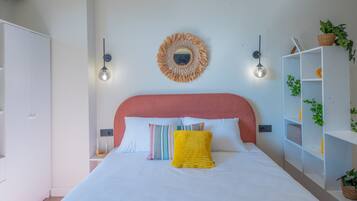 Un dormitorio con cama, cabecero, espejo fijado a la pared, dos apliques, mesita de noche y armario blanco.