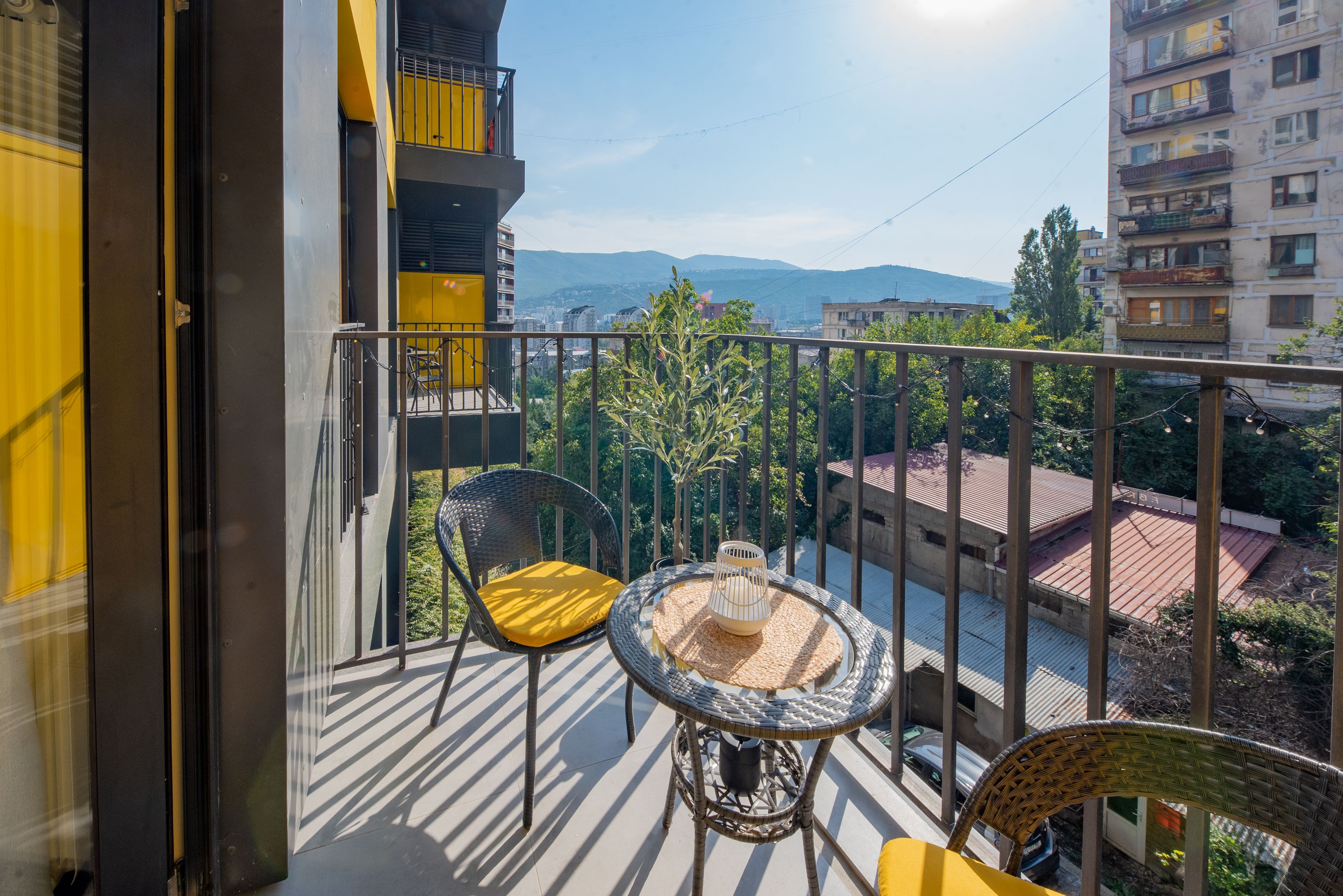A balcony with two wicker chairs and a small round table, overlooking a cityscape with buildings and mountains in the distance.