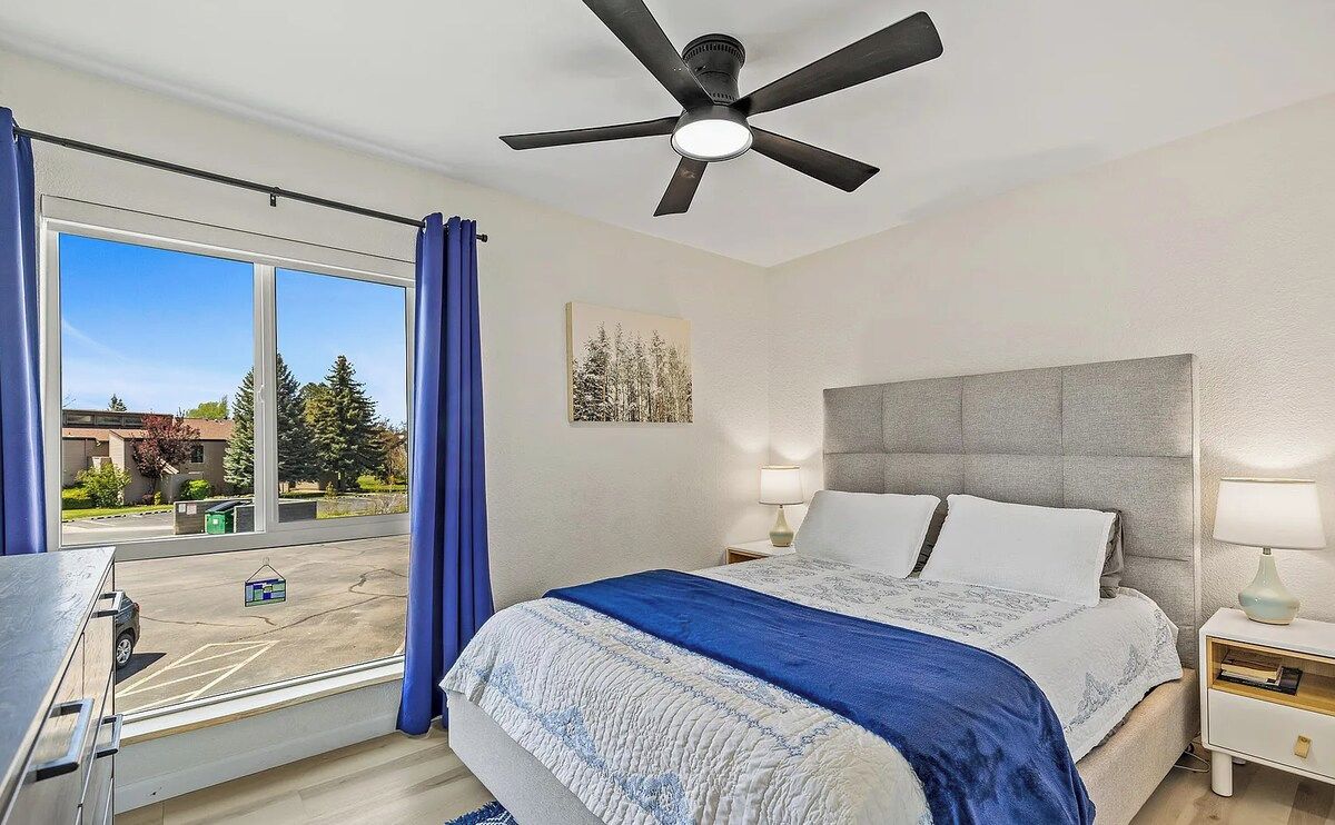 Une chambre à coucher avec un lit, un ventilateur de plafond, une table de chevet, une lampe, une fenêtre avec des rideaux et une vue sur l’extérieur.