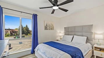 Une chambre à coucher avec un lit, un ventilateur de plafond, une table de chevet, une lampe, une fenêtre avec des rideaux et une vue sur l’extérieur.