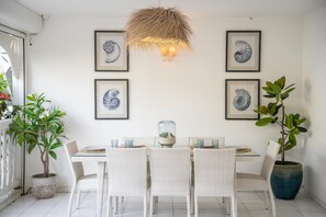 Un comedor con mesa blanca, sillas de mimbre y cuadros en la pared.