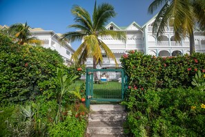 A gated entrance to a building with a white facade and a green gate, surrounded by lush greenery and a palm tree. - Savannah Élégance - Chez Soi, côté Plage (Saint-Francois)
