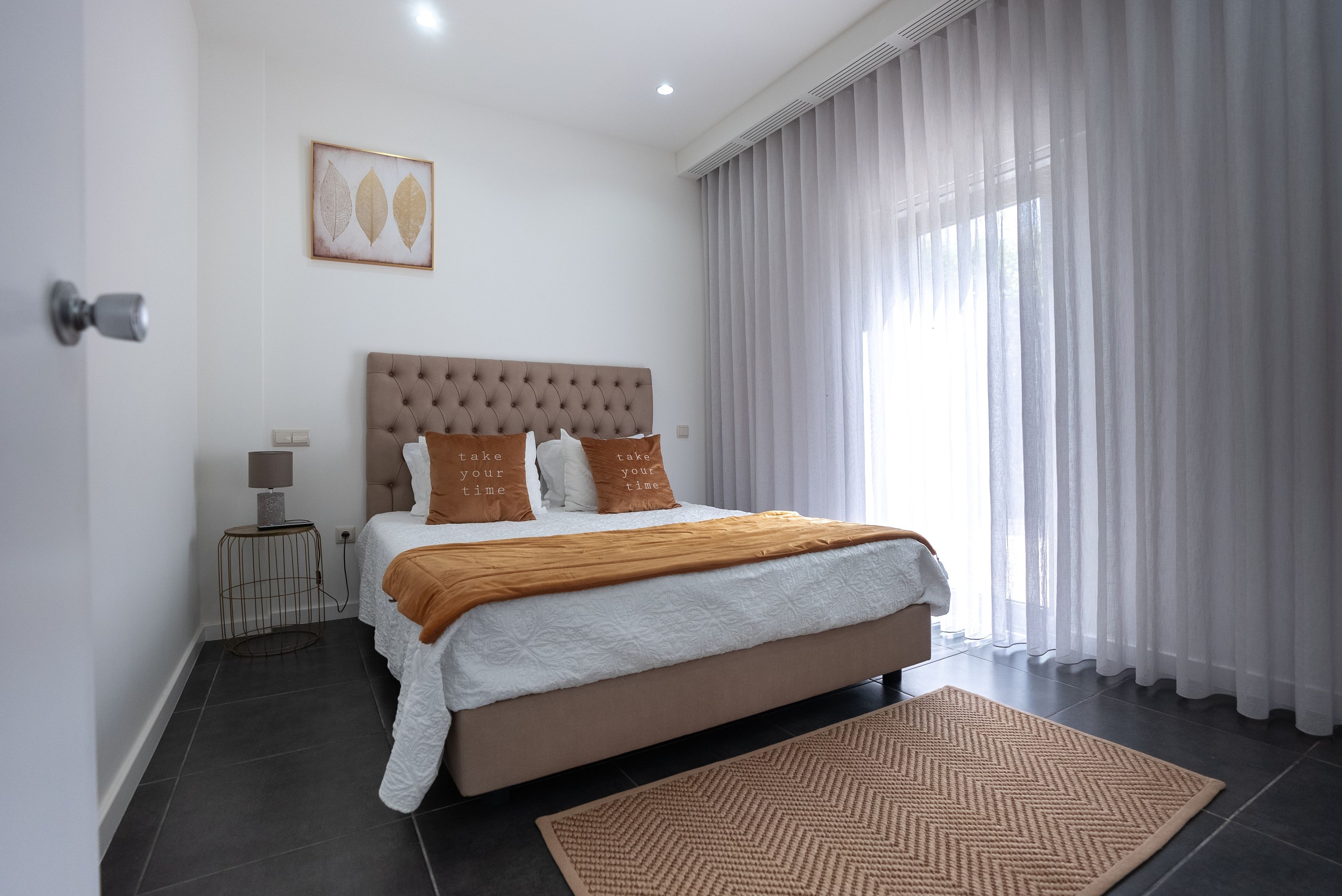 Ein modernes Schlafzimmer mit einer gepolsterten Kopfteil, einem großen Bett mit weißer Bettwäsche und braunen Kissen, einem Nachttisch mit einer Lampe und einem Blick ins Freie durch leichte Vorhänge.