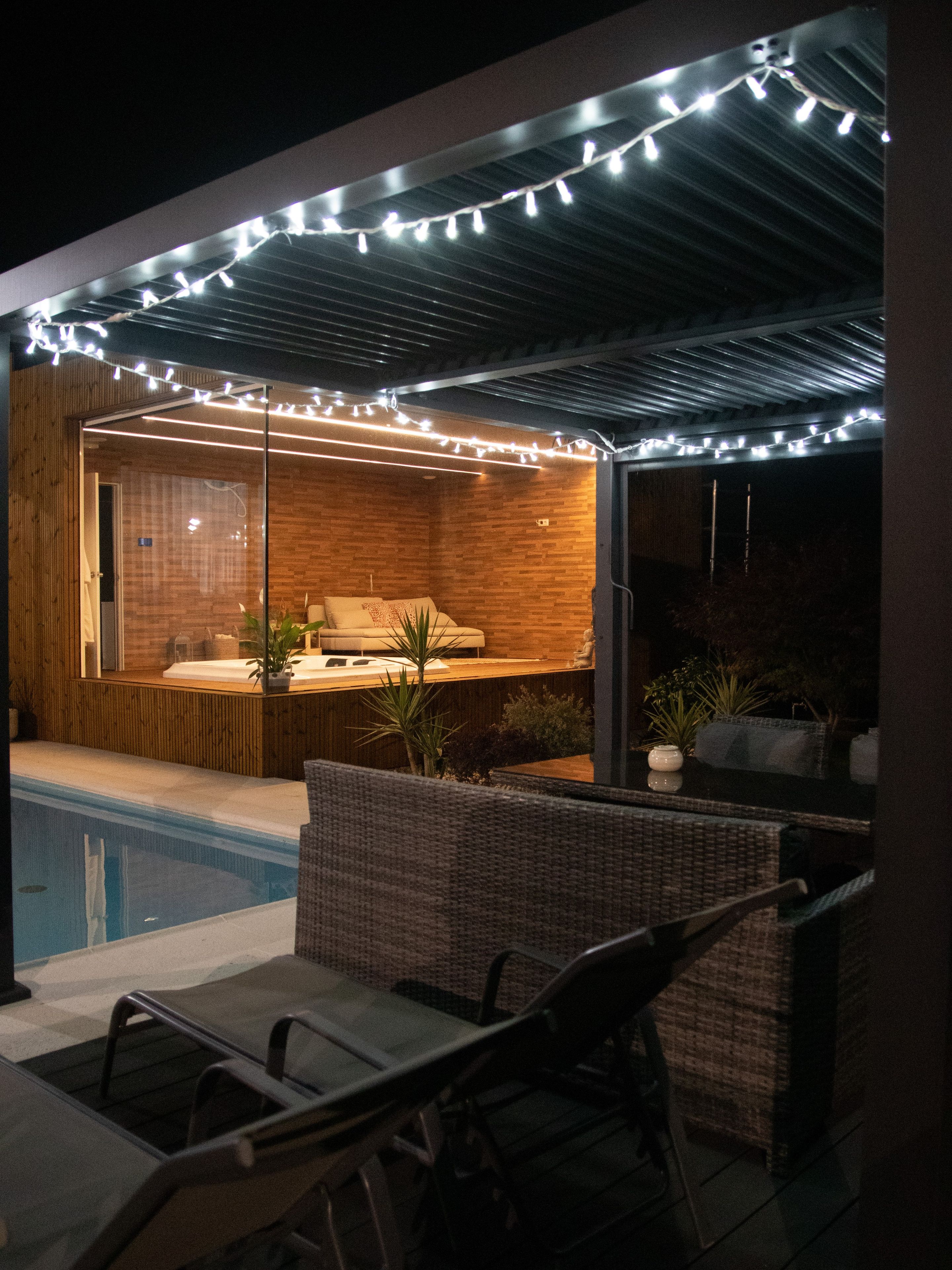 Zona de piscina con un área de estar cubierta, pared de madera y luces colgantes.