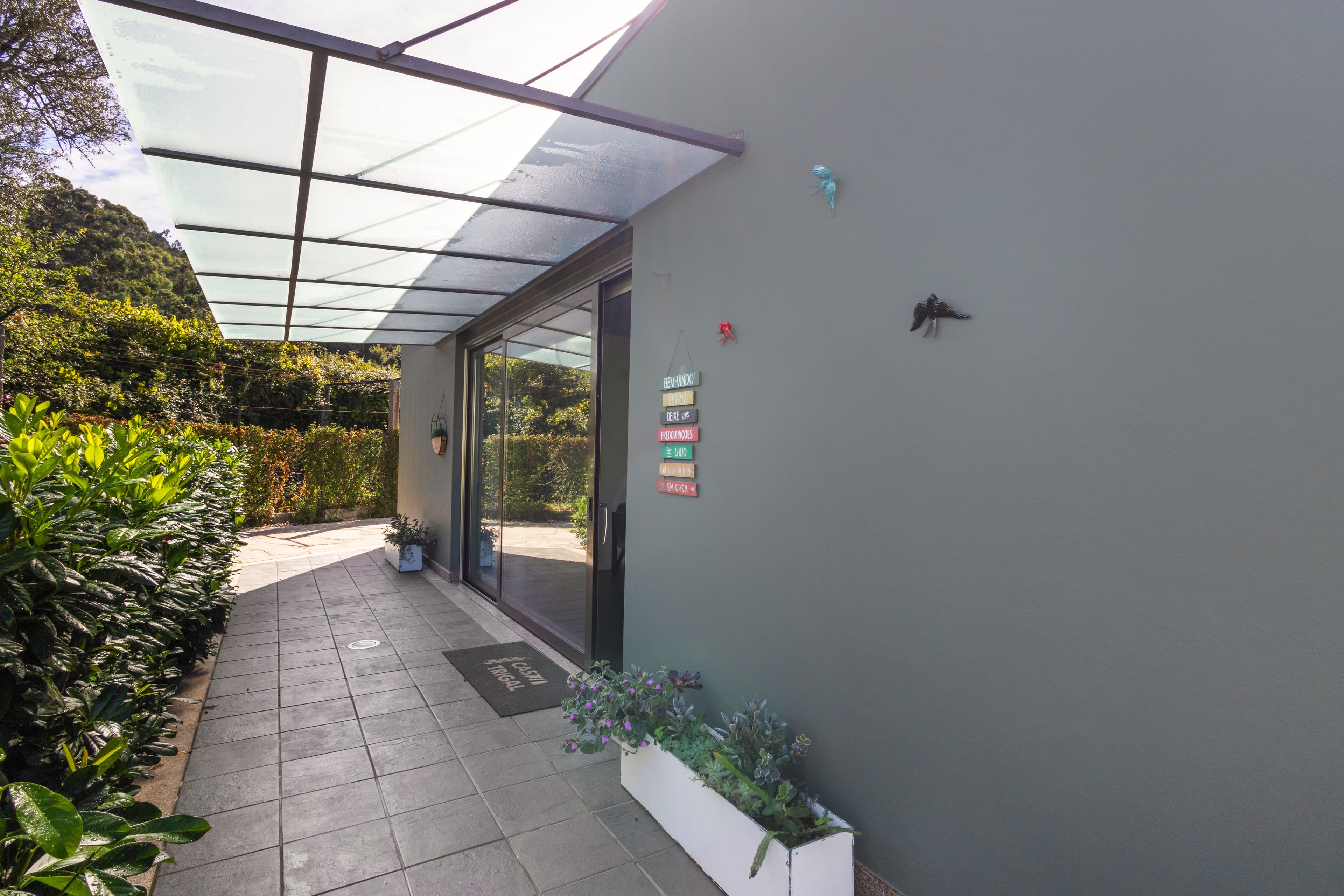 Un pasillo cubierto con techo de vidrio que conduce a la entrada de un edificio, con una puerta y un letrero en la pared que indica los nombres de las habitaciones.