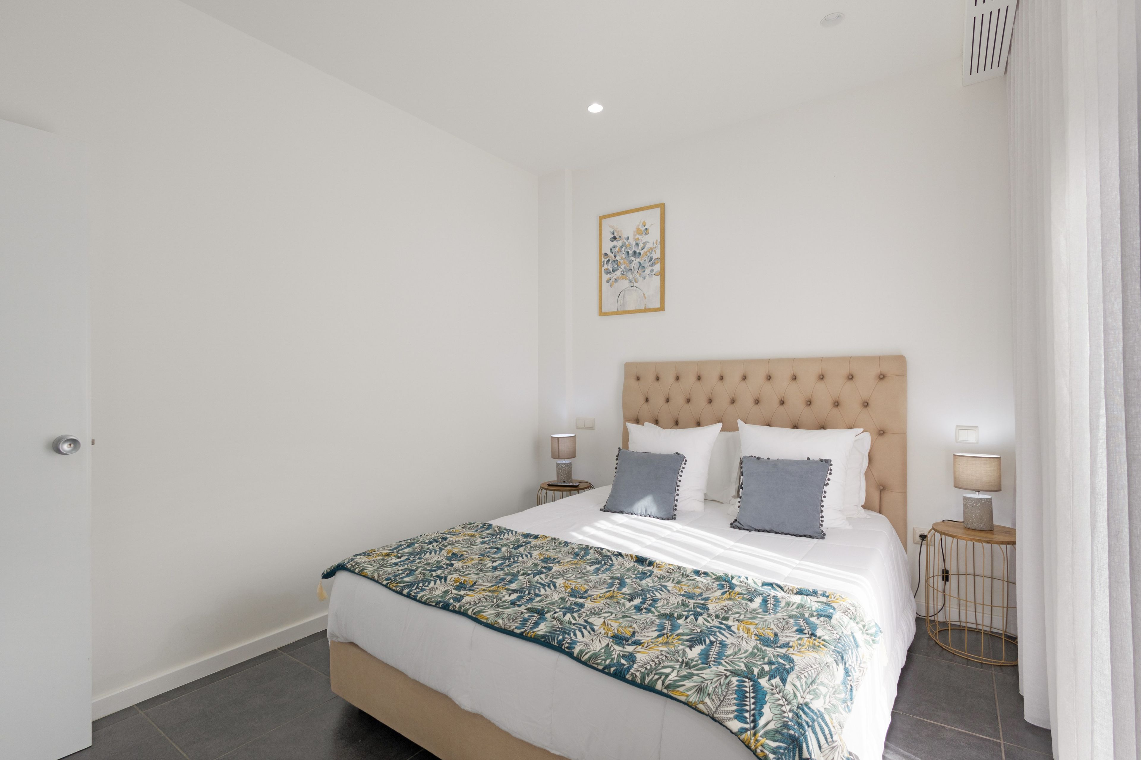A bedroom with a bed, two bedside tables, a wall-mounted light, and a framed picture.