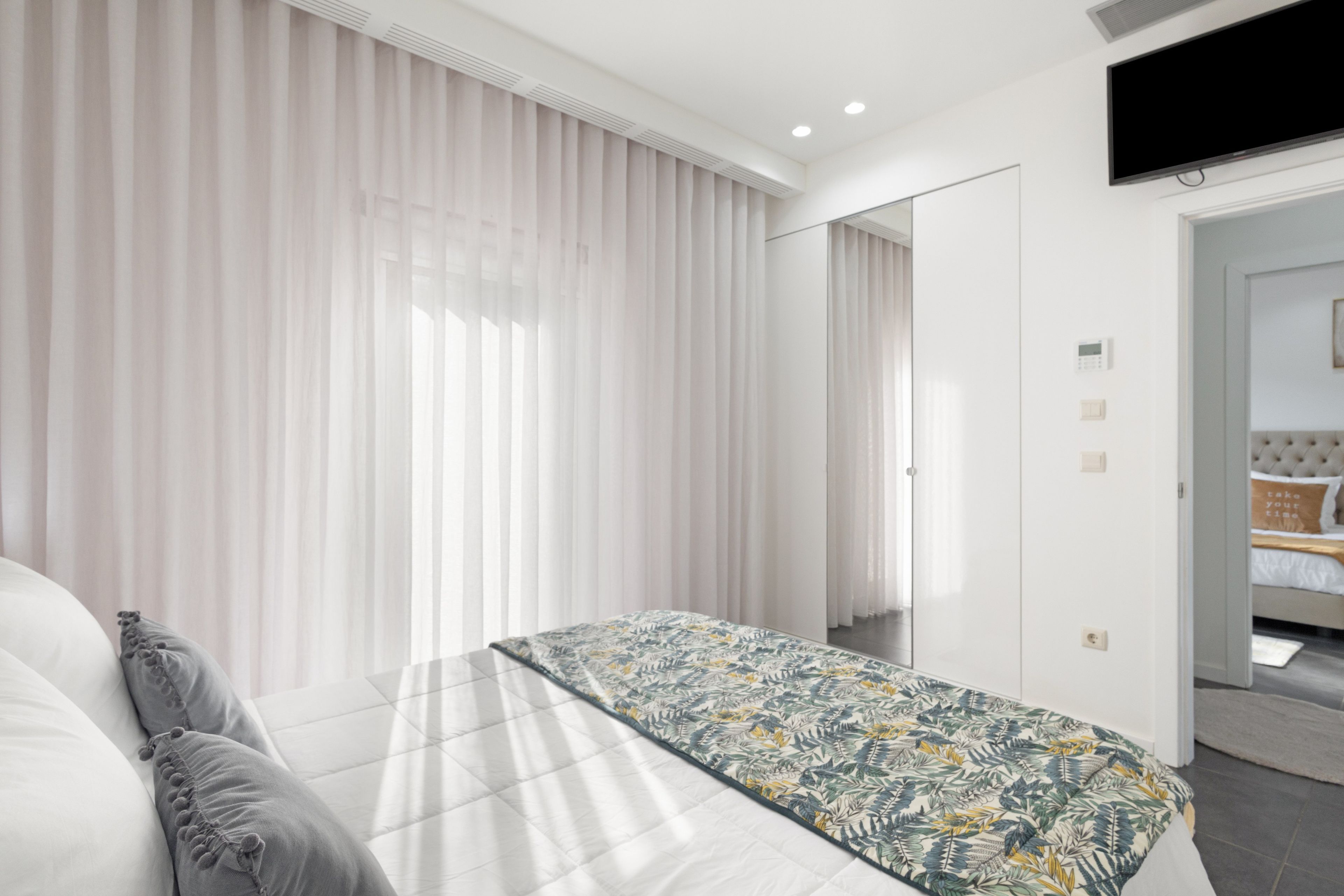 A modern bedroom with a bed, curtains, a television, and a door leading to another room.