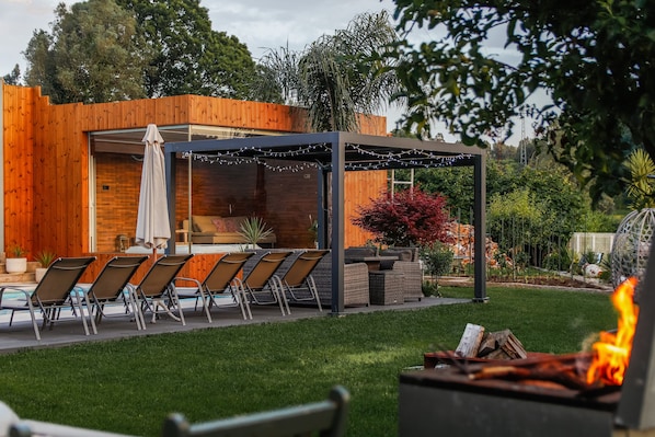 Een terras buiten met een zwembad, ligbedden en een pergola met lichtslingers.