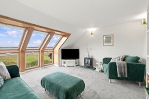 A room with a television, a sofa, a chair, and a fireplace. There are large windows and a view of the outdoors. - Schiehallion | Peaceful Scottish Retreat (Blairgowrie)